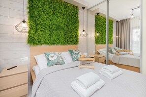 A modern hotel room with a large bed, a wooden nightstand, a small table with a cup and a book, and a green vertical garden wall. - Spacious Apartment in Gdynia | Parking | Balcony (Gdynia)