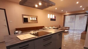 Une cuisine moderne dotée d’un îlot central, d’un système de ventilation au-dessus du plan de travail, de luminaires suspendus et d’une grande fenêtre avec stores.