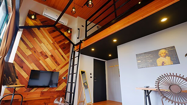 A modern interior with a wooden sloped ceiling, a mounted television, a ladder, a door, and a wall-mounted fan. - Japan Travel Accommodation (Tainan)