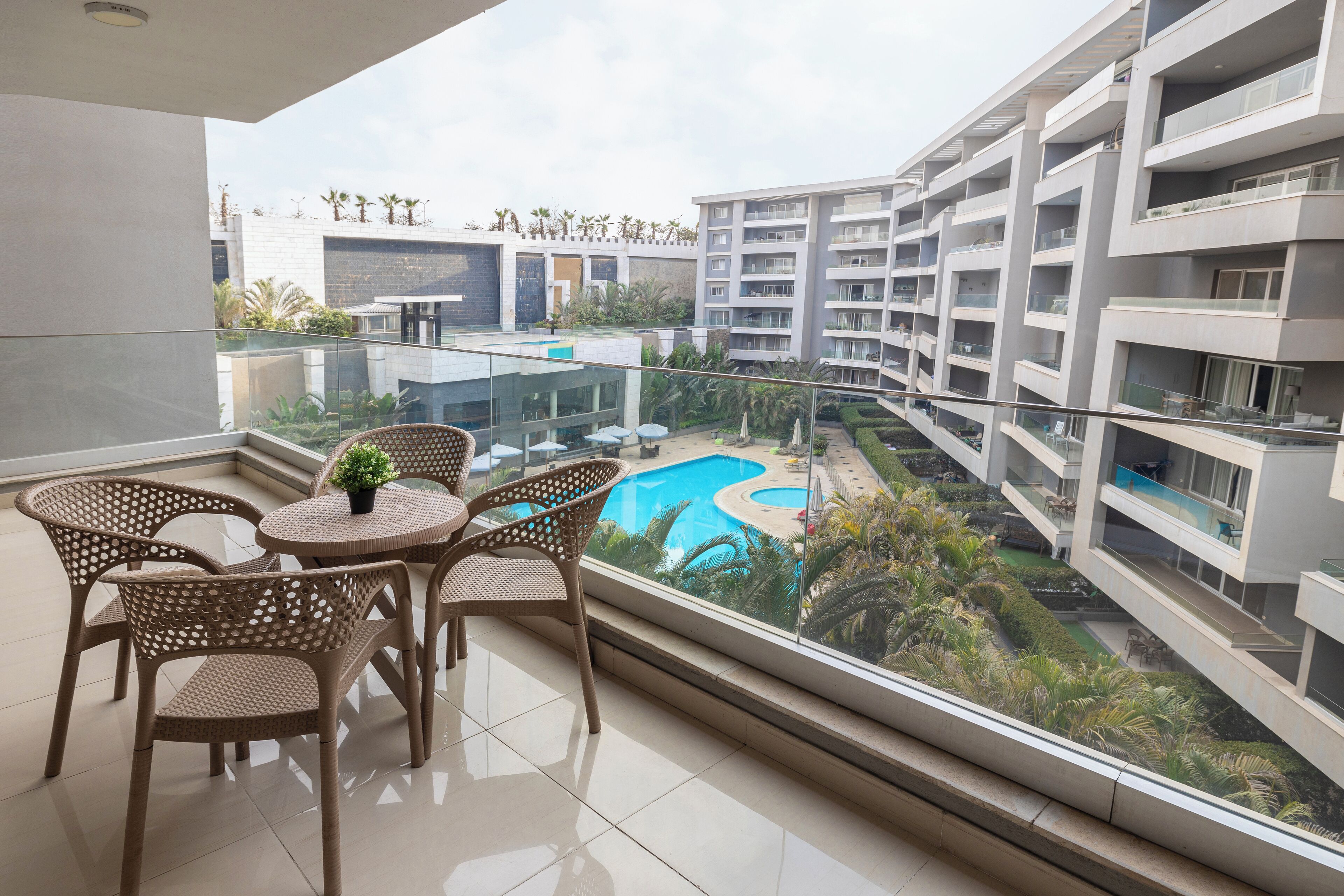 A balcony with wicker furniture, a glass railing, and a view of a swimming pool and surrounding greenery.