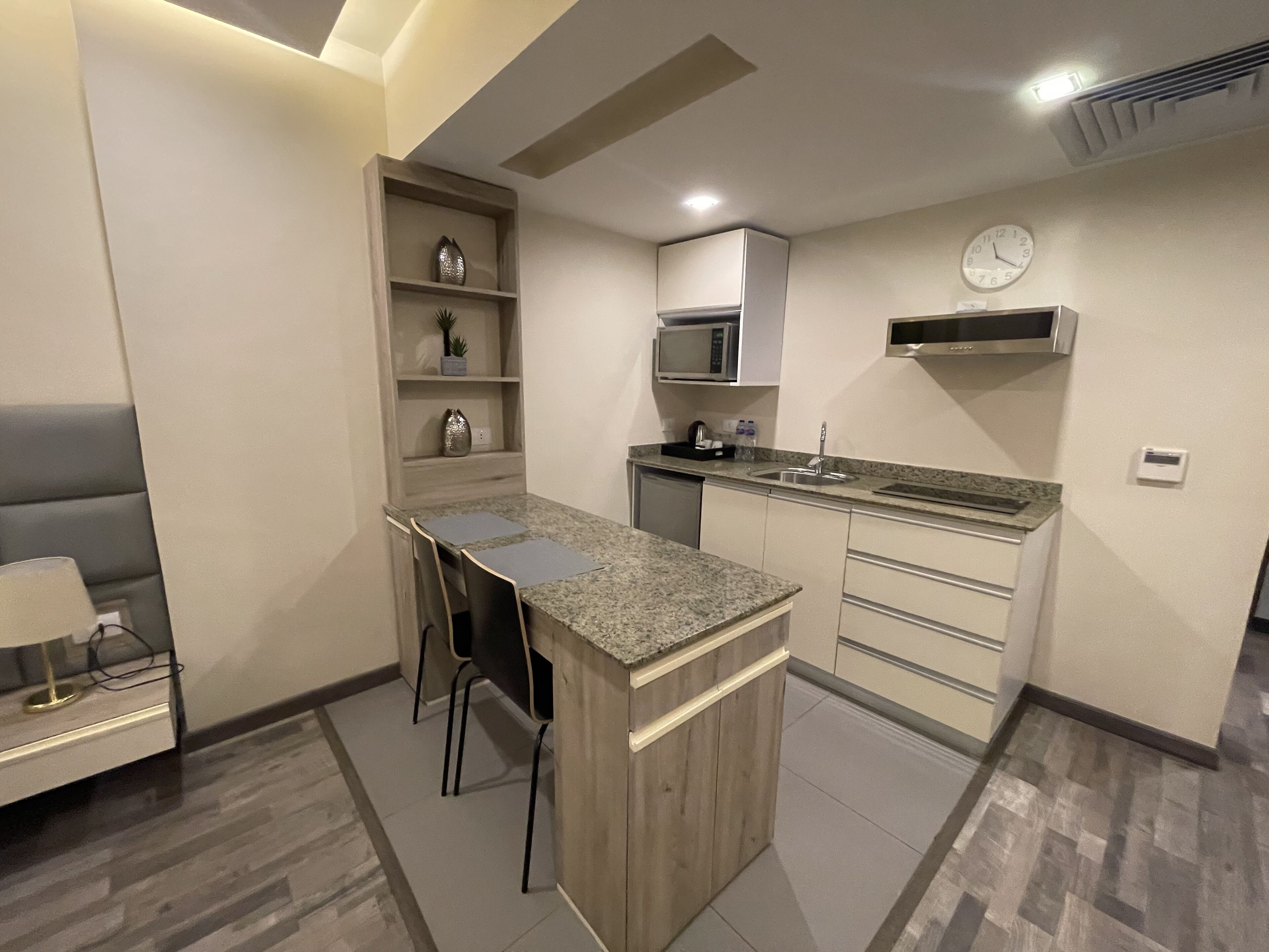 A compact kitchen with a central island, a microwave, a clock, and a small shelf with decorative items.