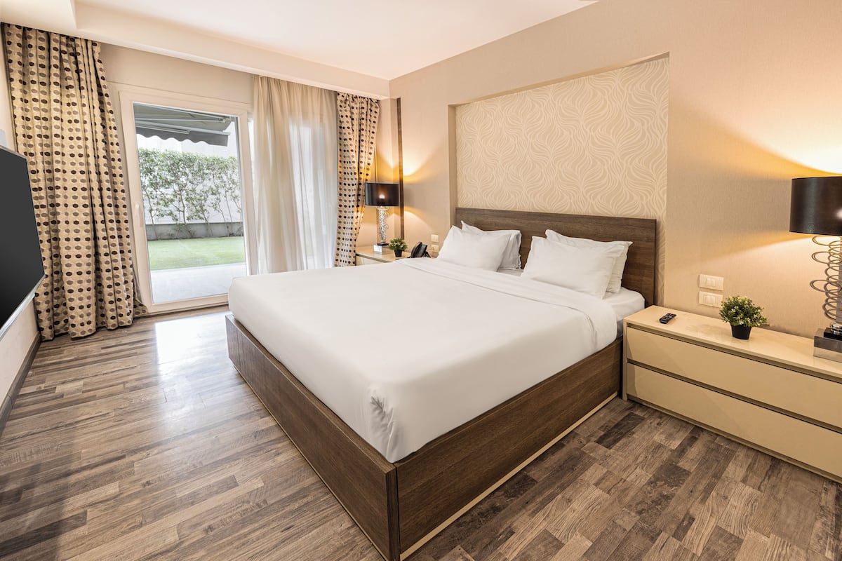 Habitación de hotel con una cama grande, suelo de madera, una mesita de noche con lámpara y vistas al exterior.