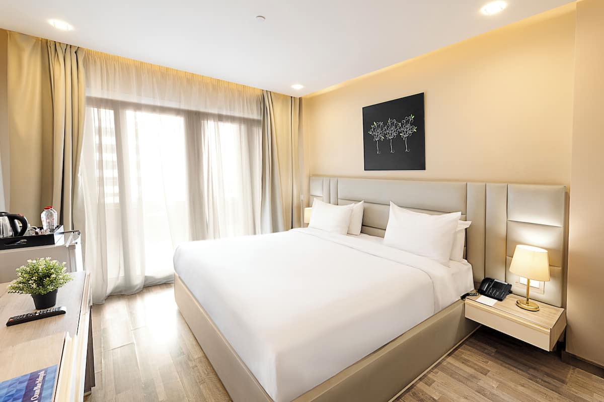 Habitación de hotel con una cama grande, mesita de noche, lámpara y vistas al exterior por la ventana.