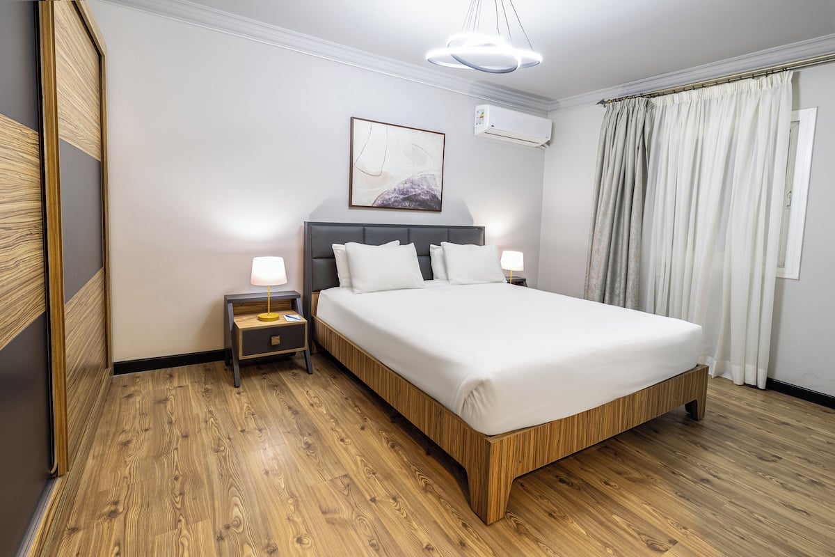 Un dormitorio ordenado con suelo de madera, una cama con ropa blanca, una mesita de noche con lámpara y aire acondicionado instalado en la pared.