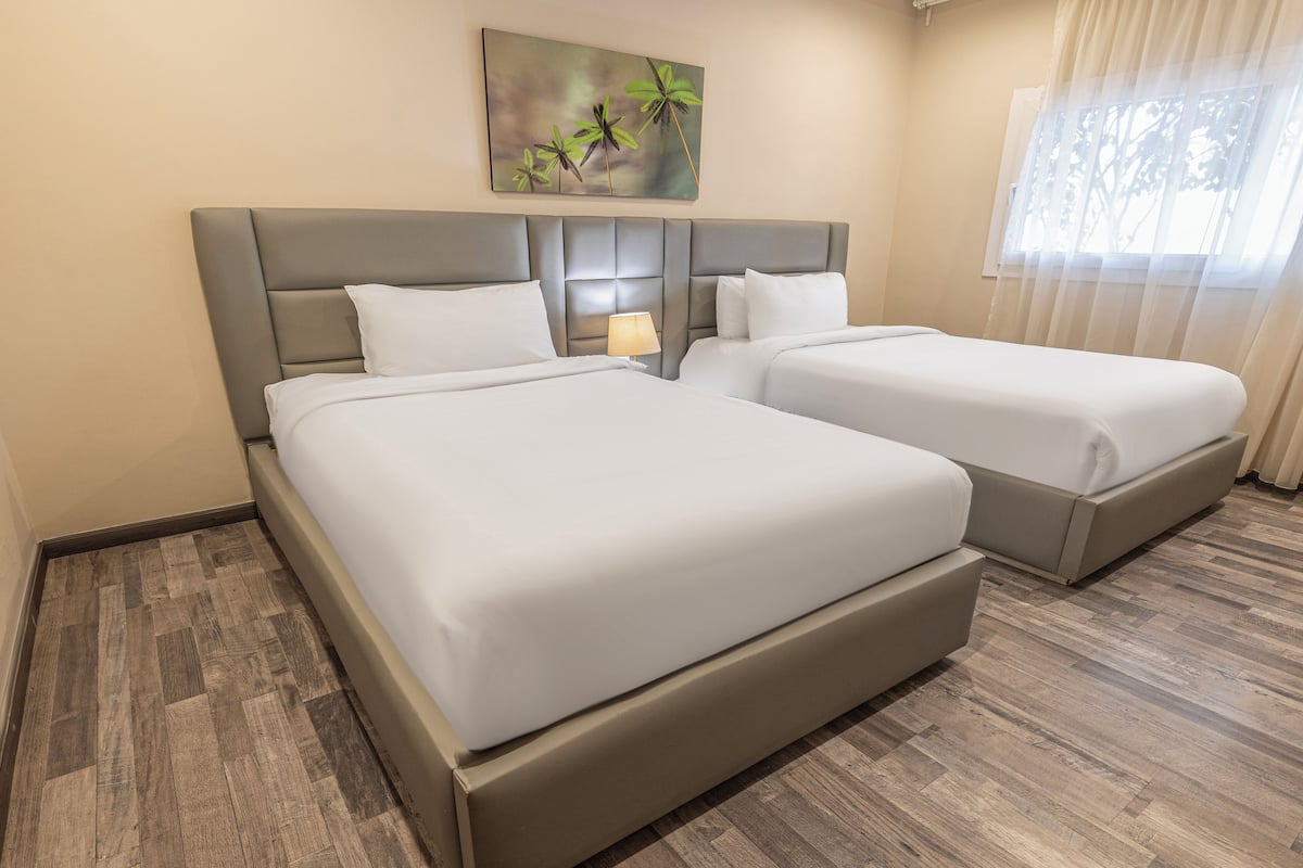 Habitación de hotel con dos camas, cabecera con lámpara y un cuadro de palmeras, además de una ventana con cortinas traslúcidas.