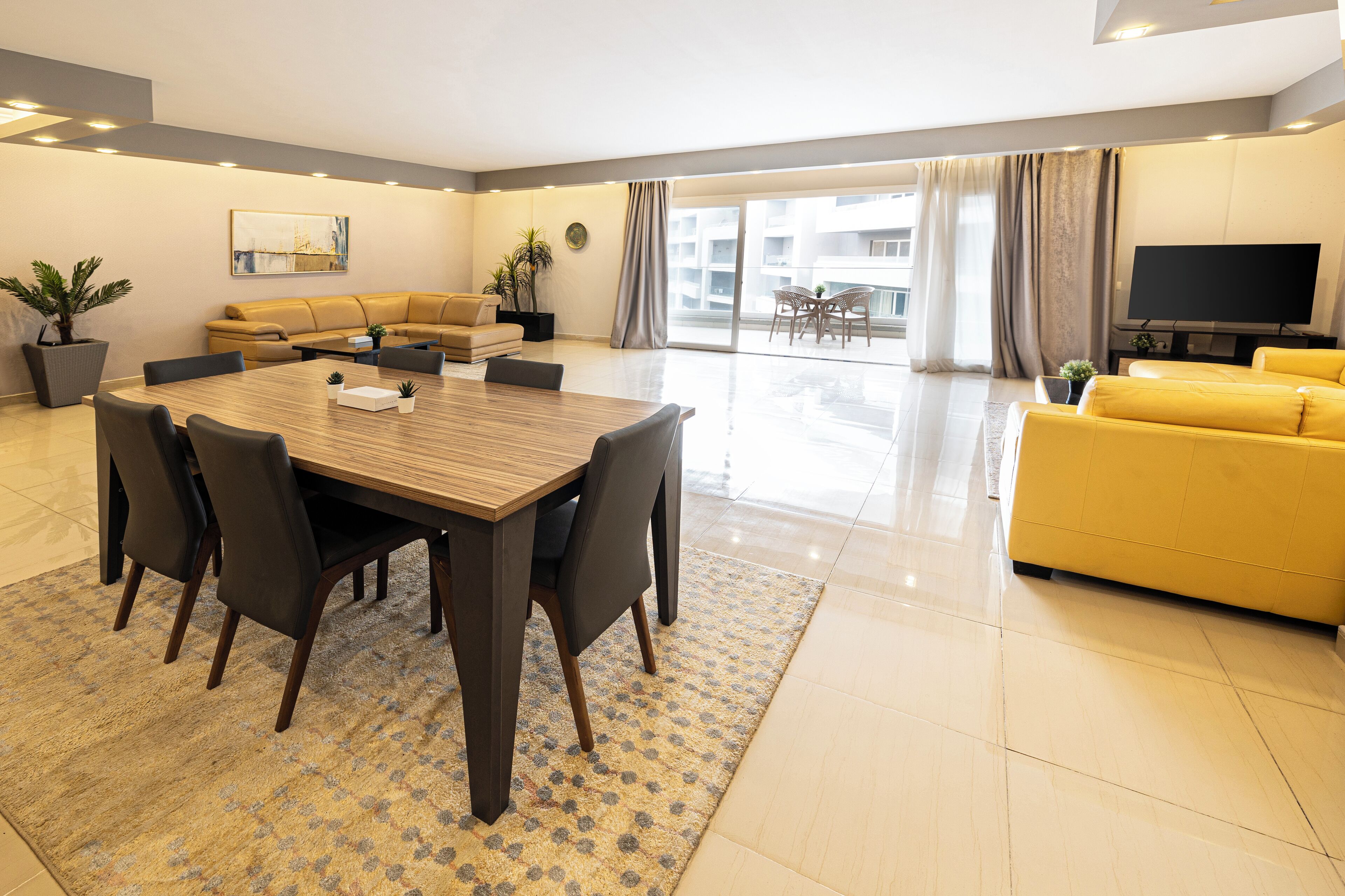 A modern living room with a wooden dining table, yellow sofa, and a flat-screen TV.