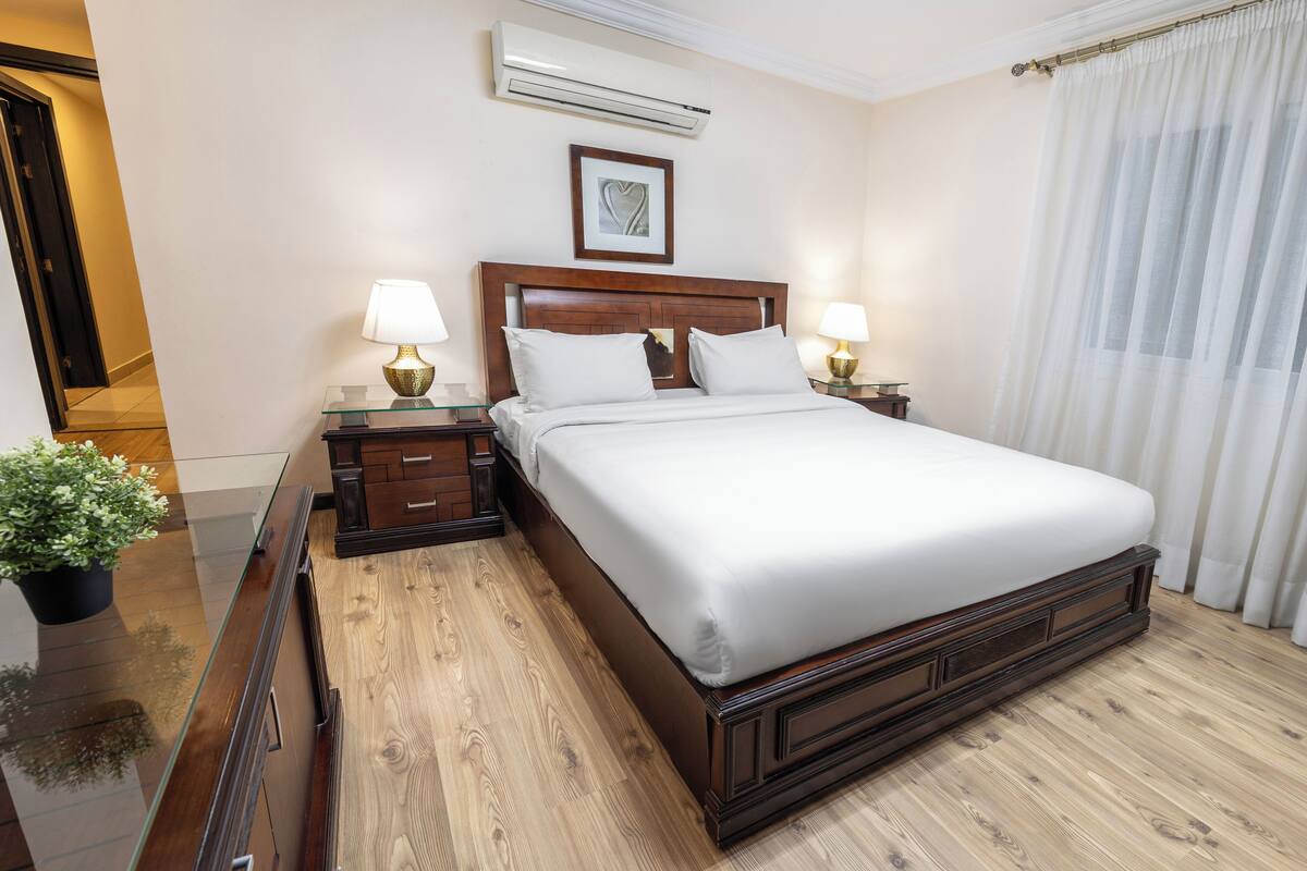 Una cama bien hecha en una habitación de hotel, con cabecera de madera, dos mesitas de noche con lámparas, una planta en maceta y un ventanal con cortinas.