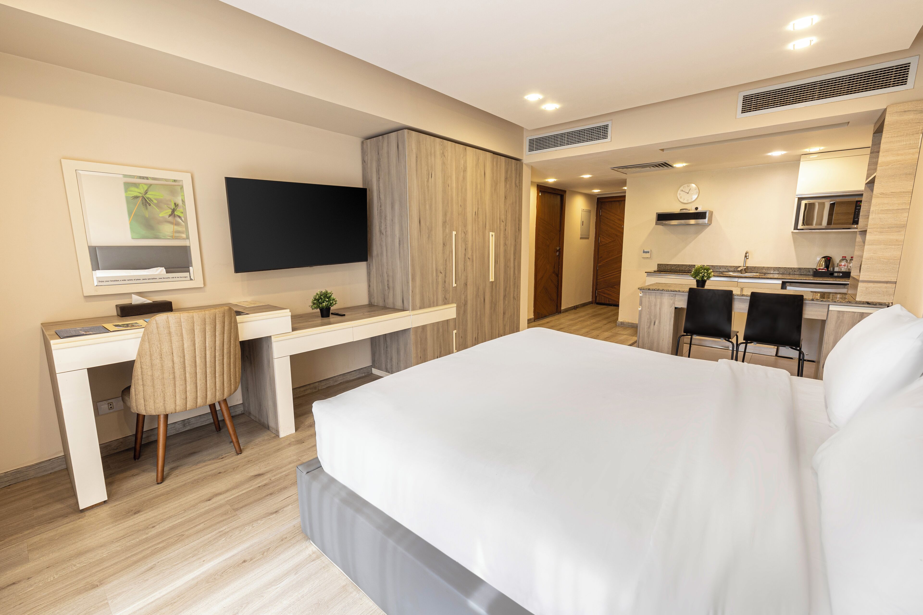 A modern hotel room with a large bed, a desk with a chair, a TV, a kitchenette, and a bathroom.