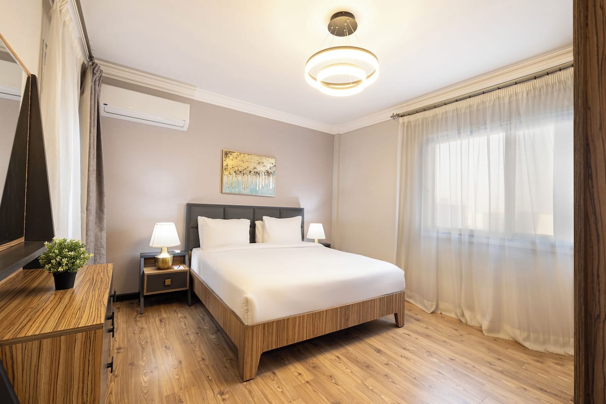 Habitación de hotel con cama, mesitas de noche, lámpara, planta en maceta, un cuadro y un gran ventanal con cortinas.