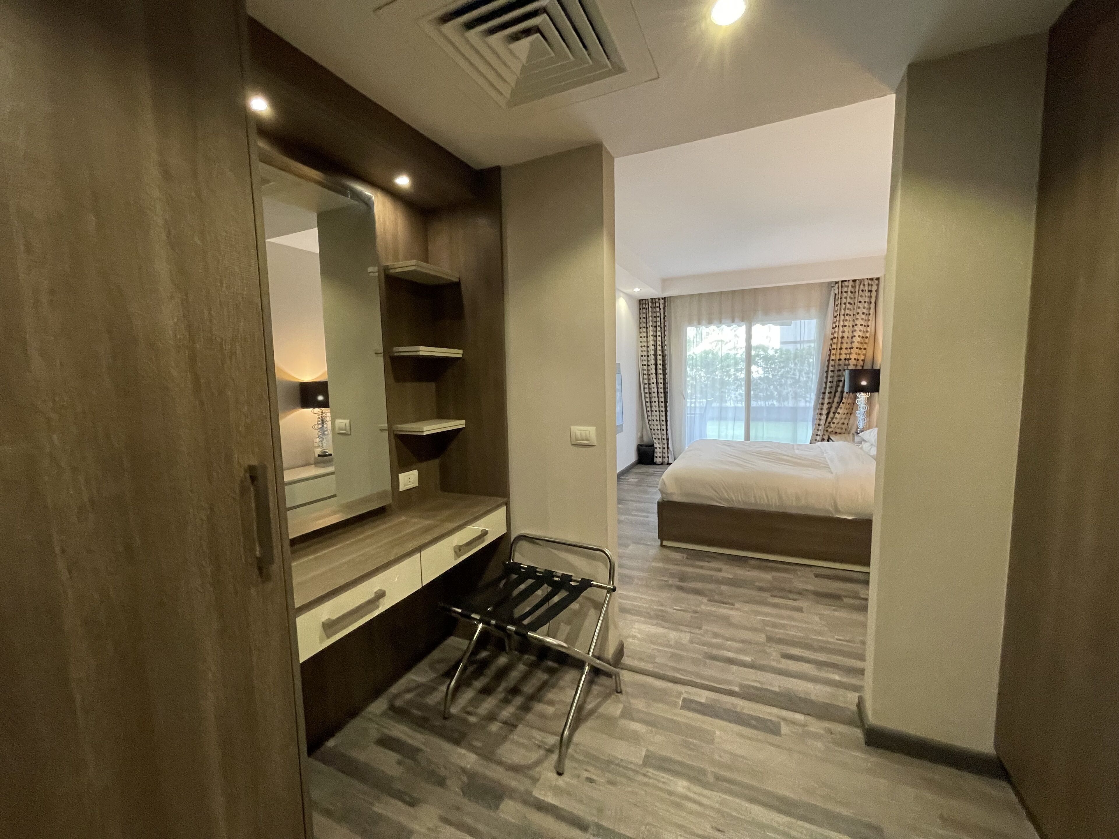 Quarto de hotel com guarda-roupa de madeira, espelho, mesa de cabeceira, cadeira preta, cama e janela com cortinas.