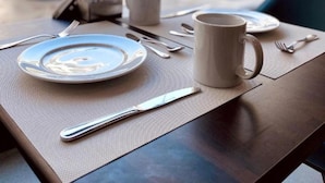A dining table with a plate, mug, and cutlery set in an outdoor setting with umbrellas and a pool in the background. - Ocean Blue Studios (Cairo)