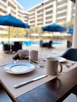 Una mesa de comedor con un plato, una taza y cubiertos, en un entorno al aire libre con sombrillas y una piscina al fondo.
