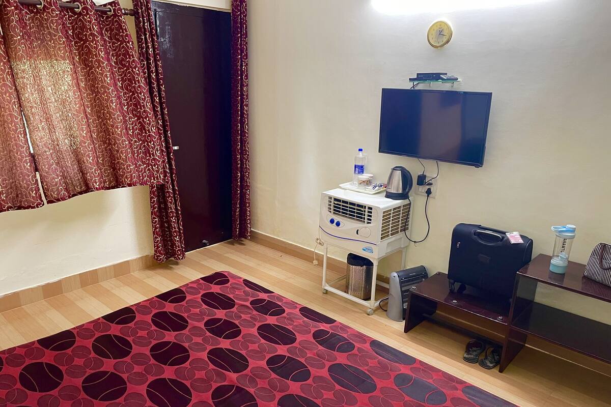 A hotel room with a TV, air conditioning unit, a small table with a bag, and a patterned rug.