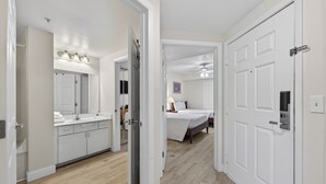 Un baño moderno con un espejo grande, lavamanos y tocador. Hay un dormitorio con cama y ventilador de techo. La habitación tiene paredes blancas y suelo de madera.