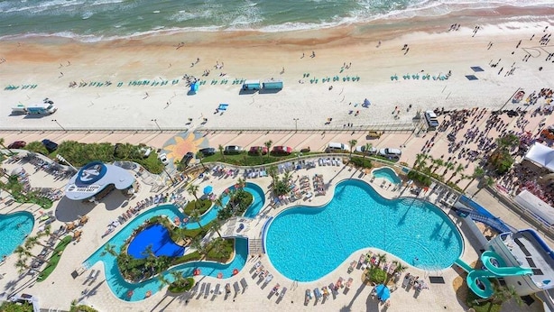 Vista aérea de un complejo turístico con una gran piscina, varias piscinas más pequeñas y una playa con gente y sombrillas.