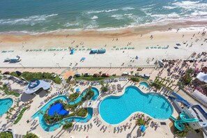 Vista aérea de un complejo turístico con una gran piscina, varias piscinas más pequeñas y una playa con gente y sombrillas.