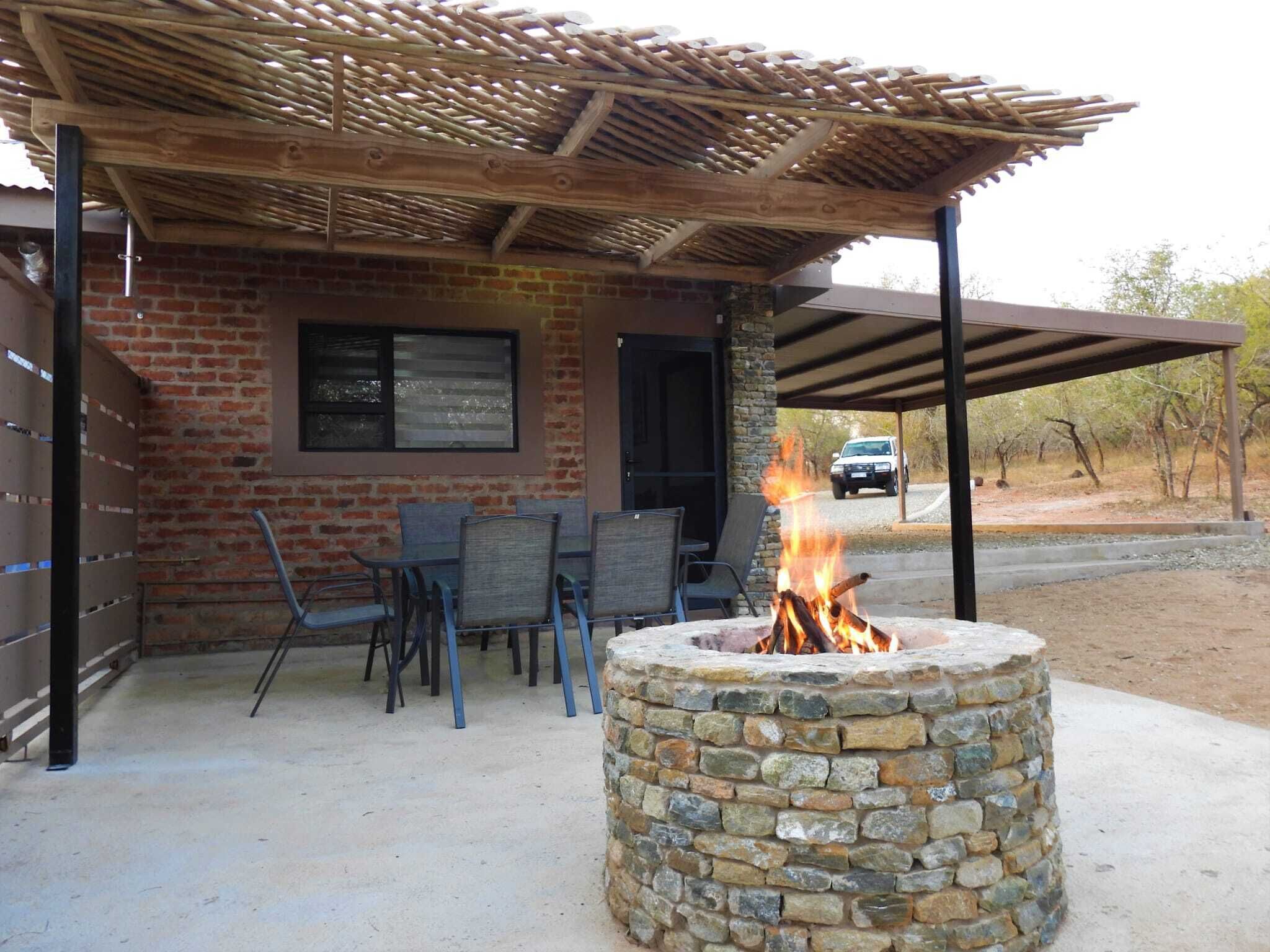 A stone fire pit under a wooden pergola, with chairs and a table nearby.