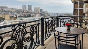 A balcony with ornate black railings, a round table with a vase of flowers, and a view of a cityscape with buildings and a body of water.
