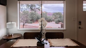 A dining area with a table, chairs, a potted plant, a lamp, and a view of a mountainous landscape through a window. - Sedona Retreat with Red Rock Views, Mid-Century Charm | Upper Level Duplex (Sedona)