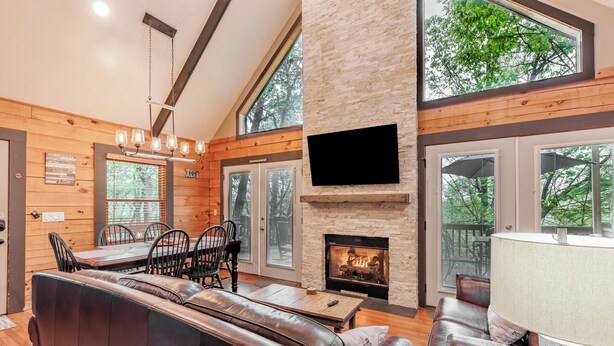 A spacious living area with a fireplace, leather seating, a dining table with chairs, and large windows offering a view of trees.