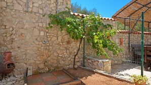Um muro de pedra com um portão de metal, uma pequena área de jardim com um banco de metal e um edifício rústico com telhado de telhas.