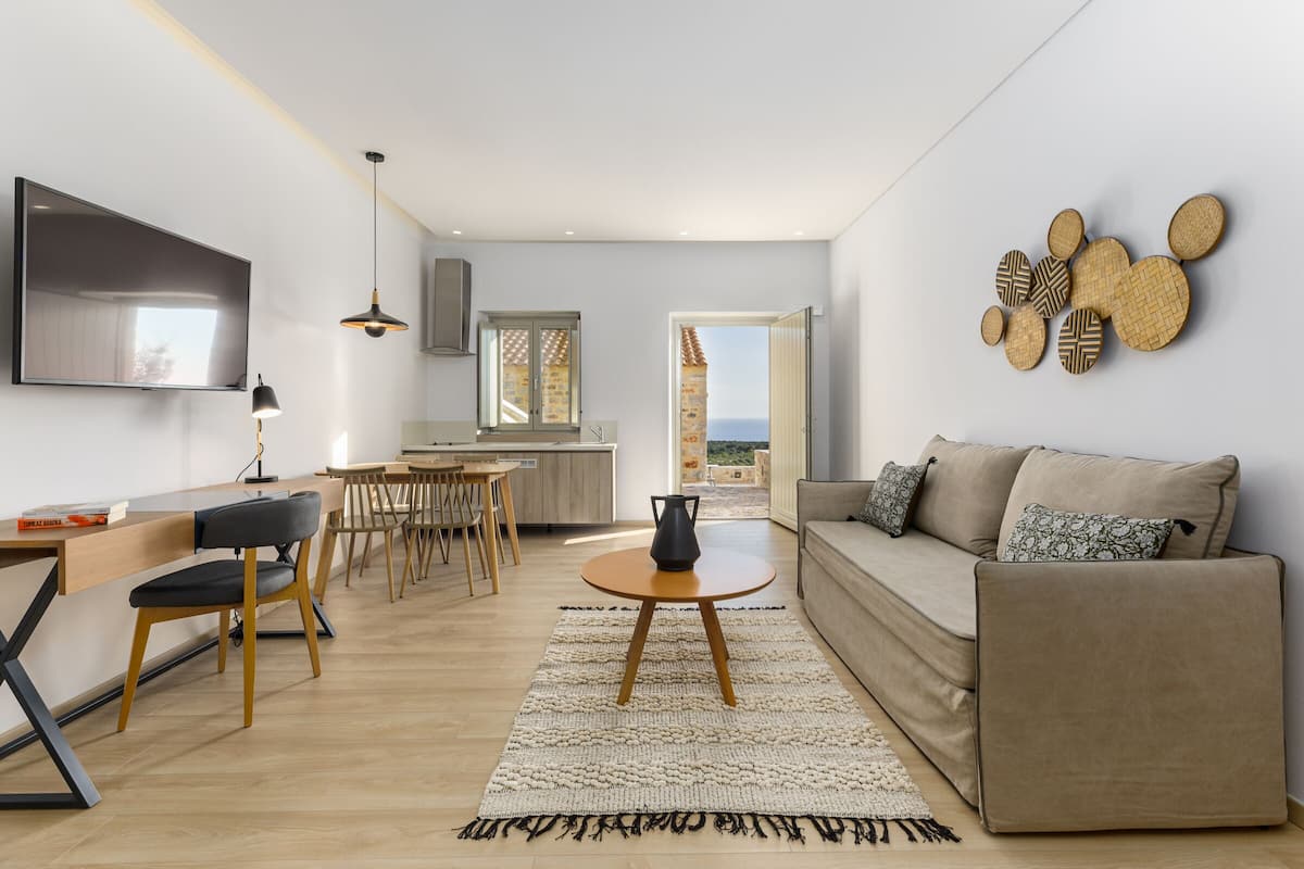 Una sala de estar moderna con sofá, mesa de centro y televisión. Se ve una cocina al fondo, con vista al mar.
