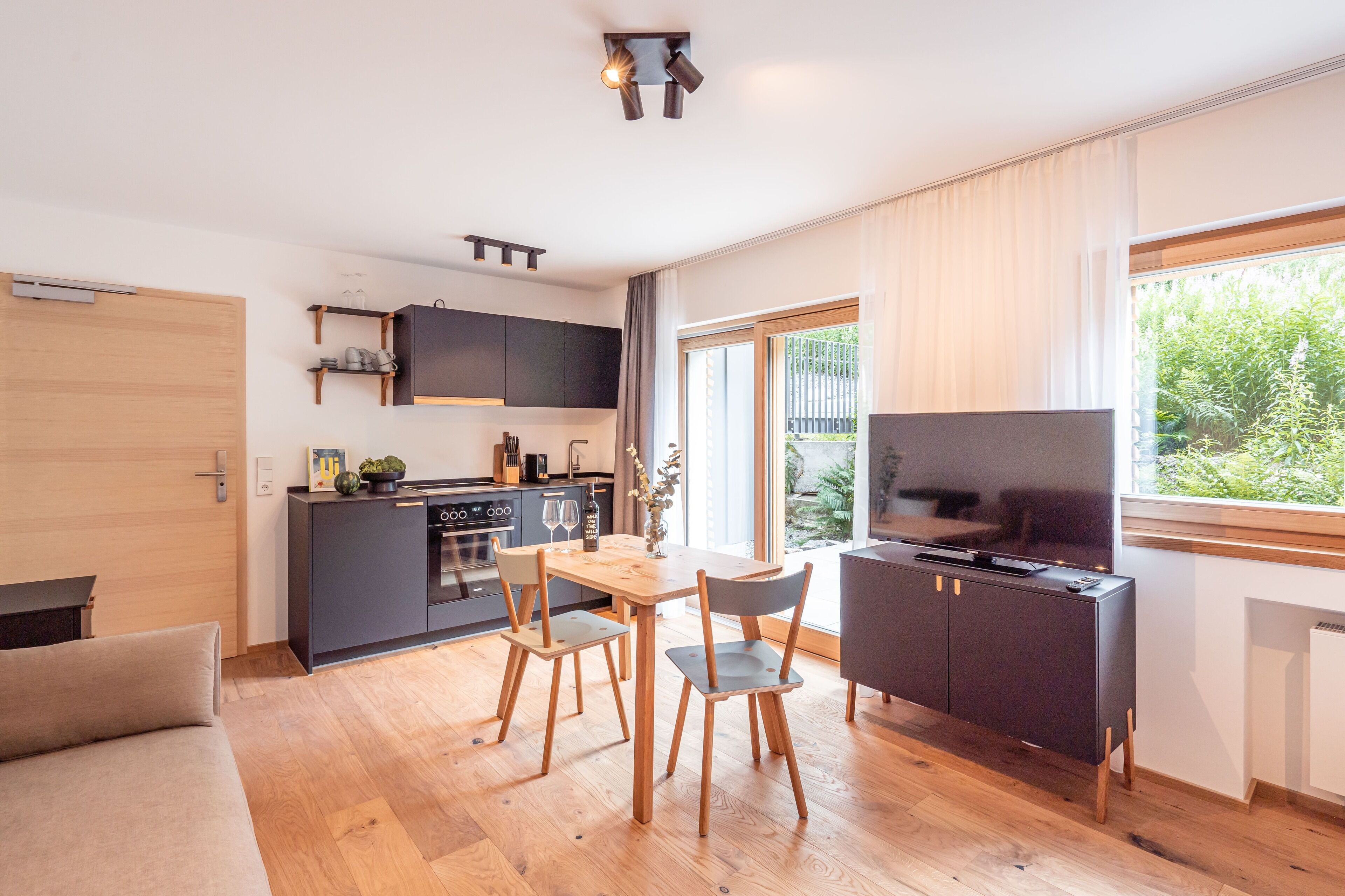 Eine moderne Küche mit Holzschränken, einem Esstisch mit Stühlen, einem Fernseher und einem großen Fenster mit Blick ins Grüne.