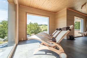 A modern interior with wooden ceiling and walls, large windows offering a view of a green landscape, and several wooden lounge chairs. - stuub todtnauberg (Todtnau)