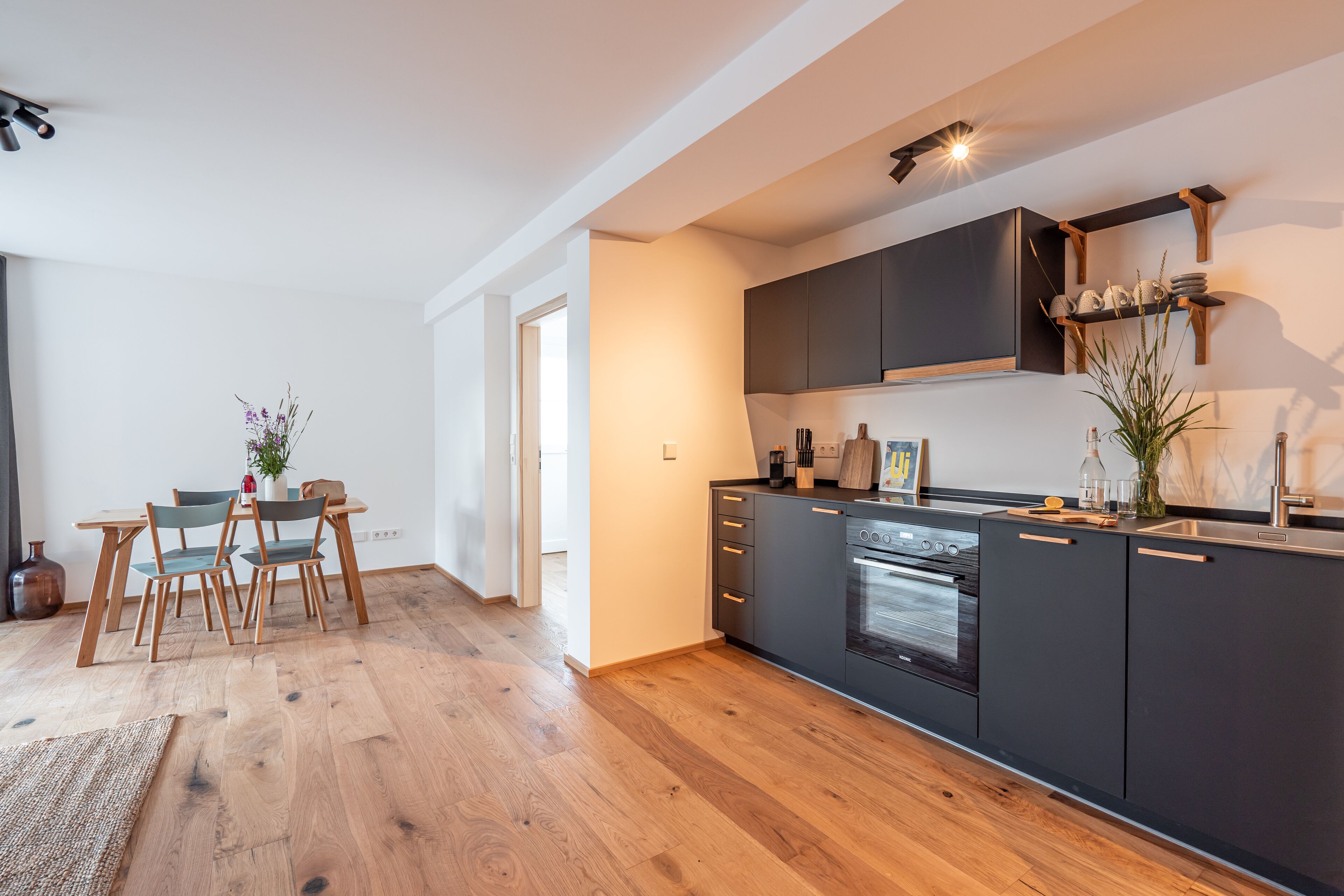 A modern kitchen with dark cabinets, a built-in oven, and a dining area with wooden chairs and a table.