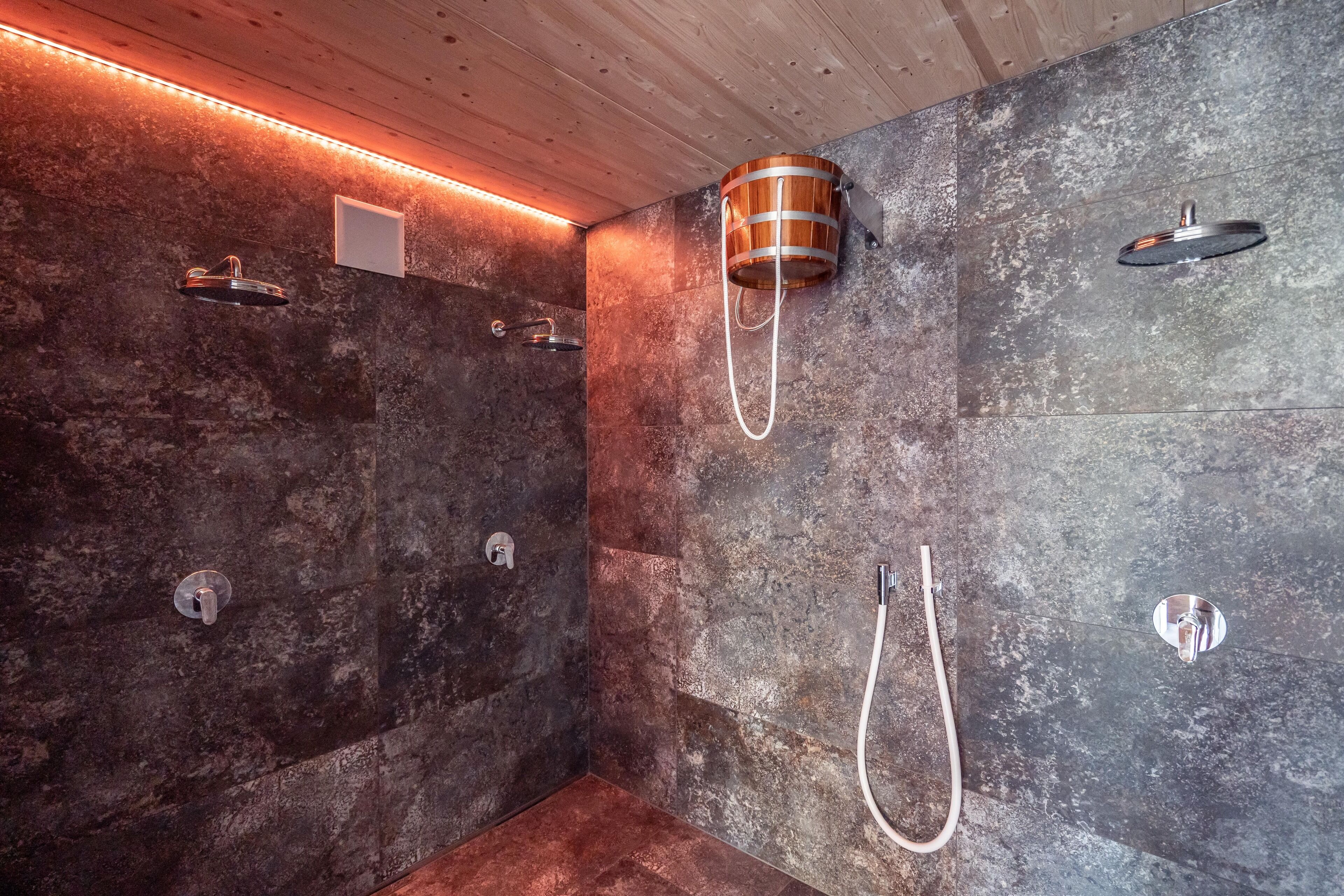 Un área de ducha moderna con paredes de piedra y detalles de madera, que incluye múltiples cabezales de ducha y un original cabezal de ducha en forma de cubo de madera.