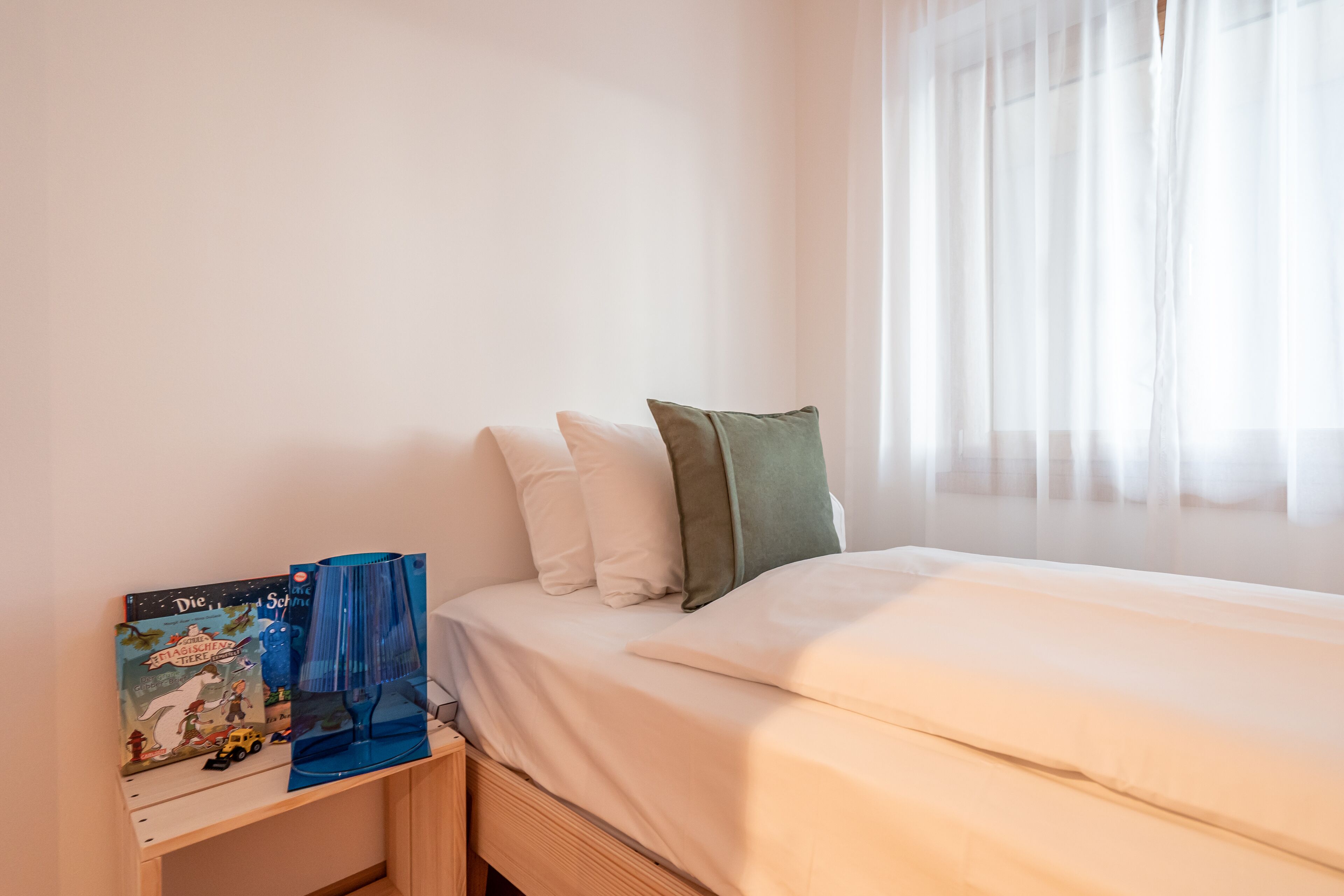 Une chambre à coucher avec un lit, des oreillers, une table de chevet avec un livre, et une fenêtre avec des rideaux légers.