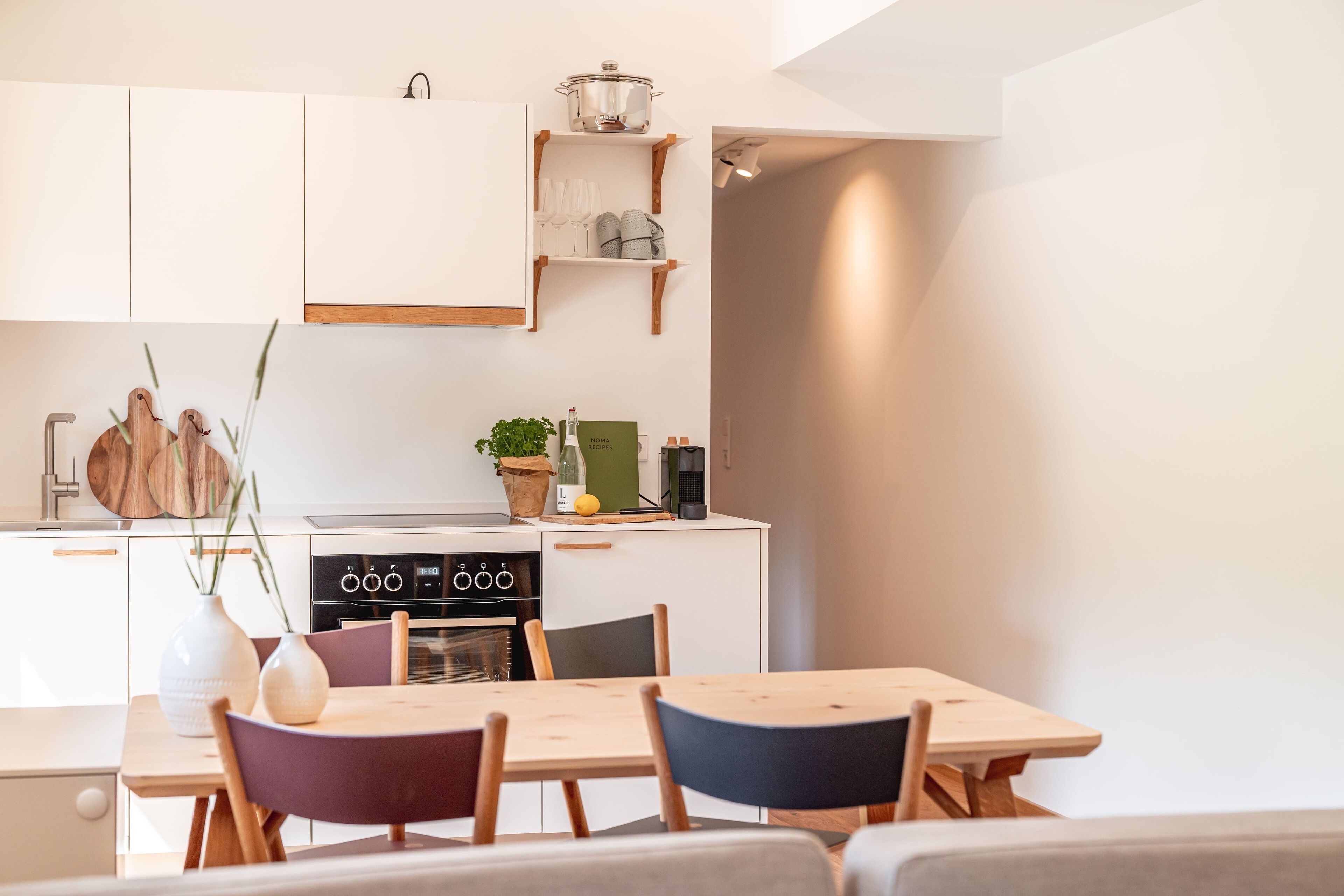 Una cocina moderna con armarios blancos, horno empotrado y zona de comedor con mesa y sillas de madera.