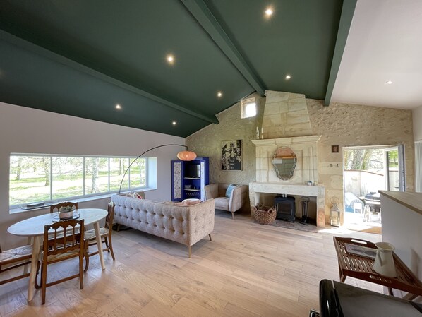 A spacious living room with a fireplace, a dining area with a round table and chairs, and a large window offering a view of the outdoors.