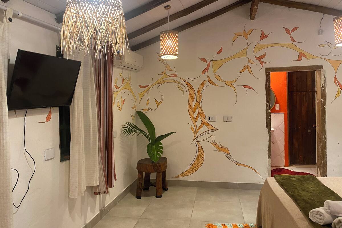 Une pièce avec un téléviseur fixé au mur, une fresque murale représentant un arbre décoratif, un petit tabouret en bois avec une plante, et un lit recouvert d’une couverture verte.