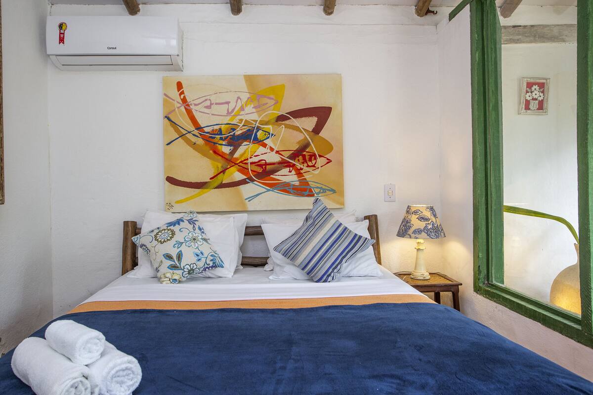 Un lit bien fait, recouvert d’une couvre-lit bleue, avec des oreillers aux motifs floraux et rayés, et une peinture abstraite aux couleurs vives accrochée au mur.