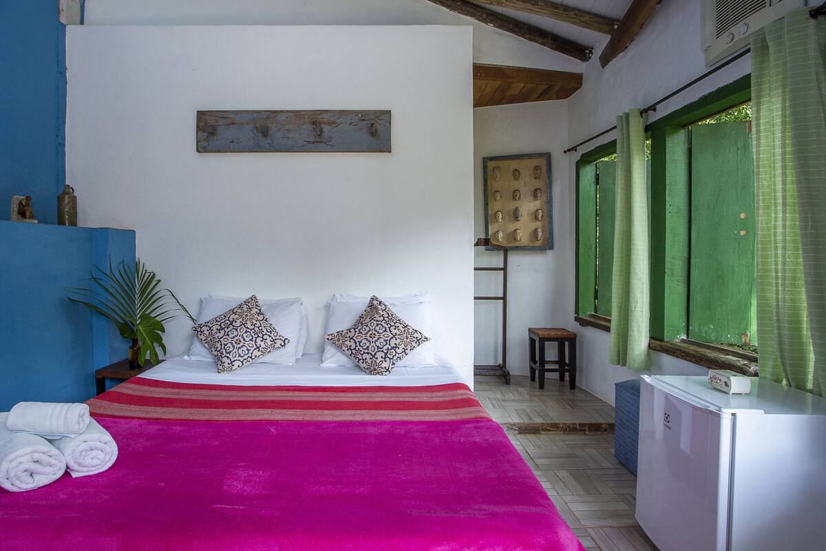 Une chambre à coucher avec un lit, des oreillers, une couverture, une petite table, une plante et une fenêtre avec des rideaux.