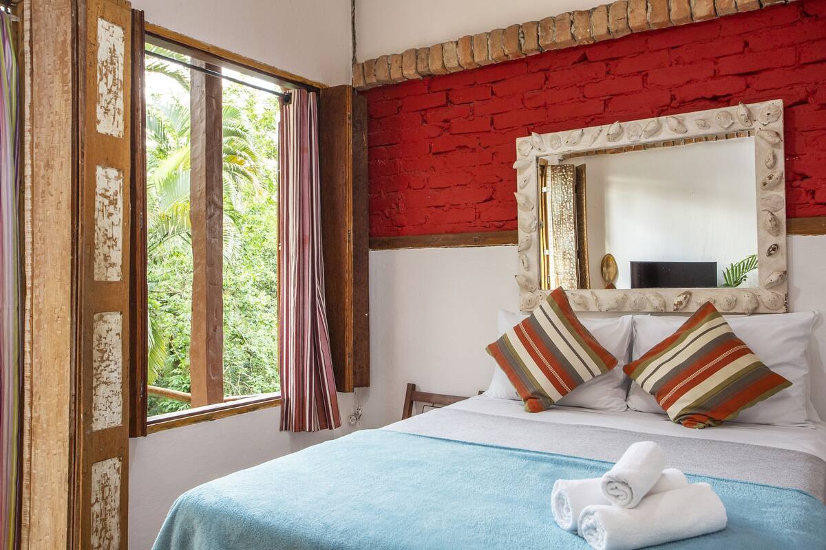 Une chambre d’hôtel avec un lit, une fenêtre avec des rideaux, un miroir et une vue sur la verdure.