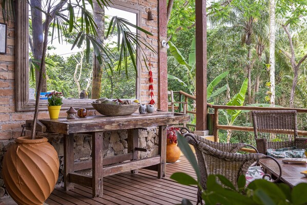 A rustic wooden table with a potted plant, a bowl, and a teapot on a deck with wicker chairs.