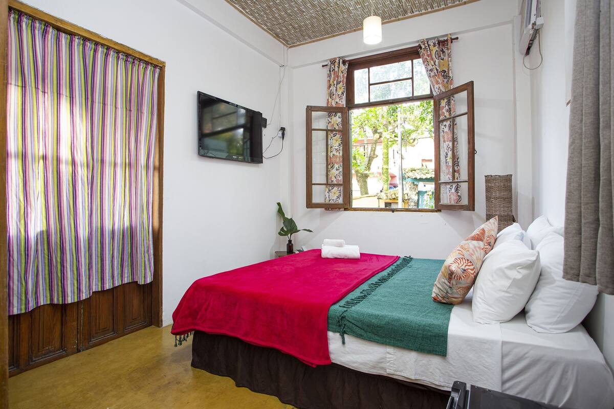 Une chambre avec un lit, une télévision, une fenêtre avec des rideaux et un plancher en bois.