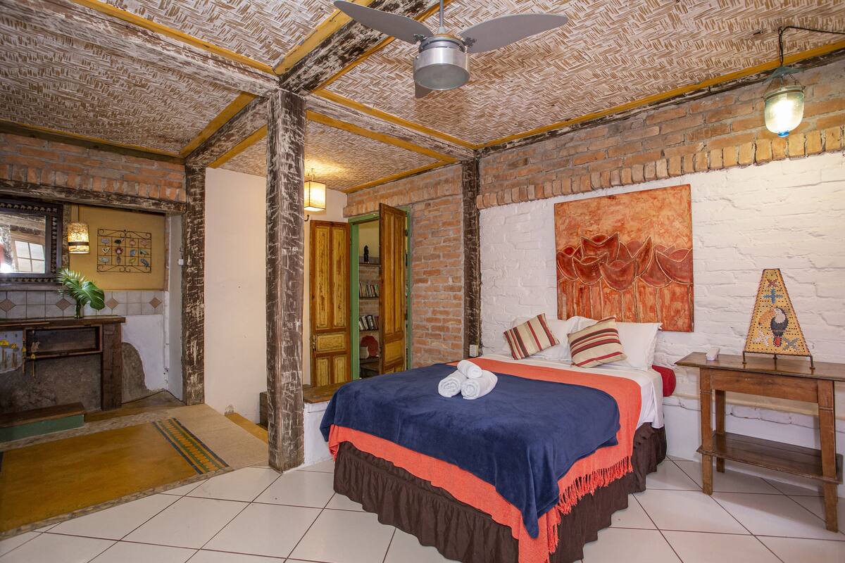 Une chambre avec un mur en briques, un lit recouvert d’une couverture bleue et rouge, une table de chevet en bois et un ventilateur de plafond.