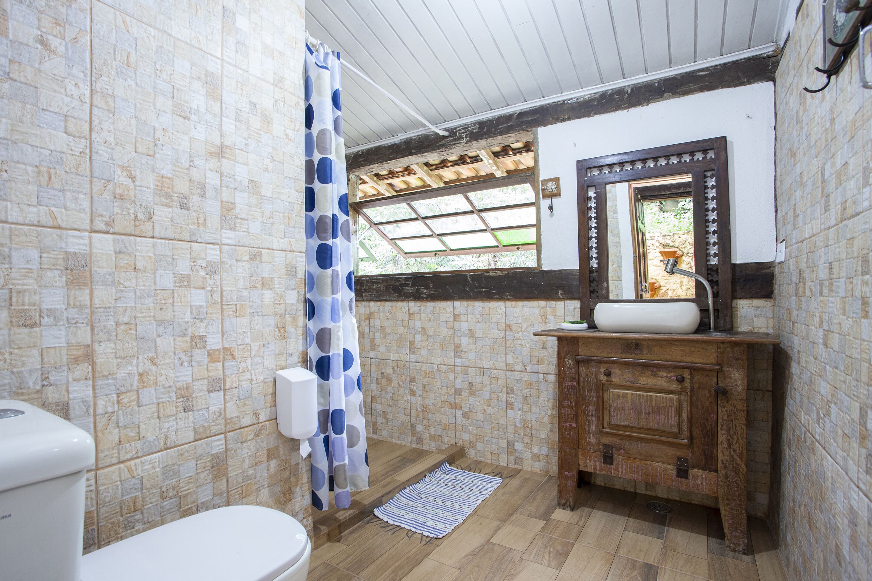 A bathroom with a toilet, shower, wooden cabinet, and a window with a view of greenery.