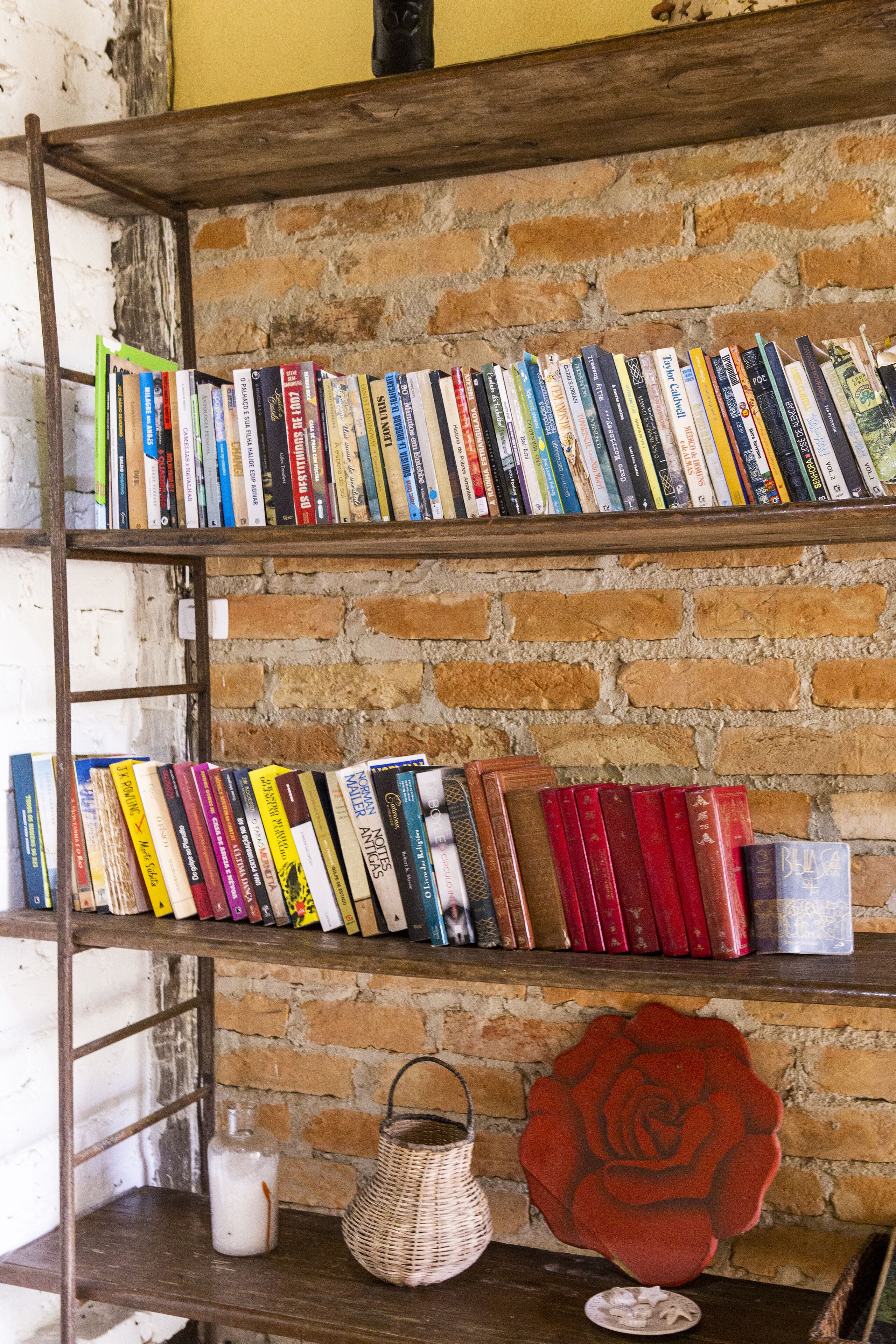 A bookshelf against a brick wall with a variety of books and a decorative rose.