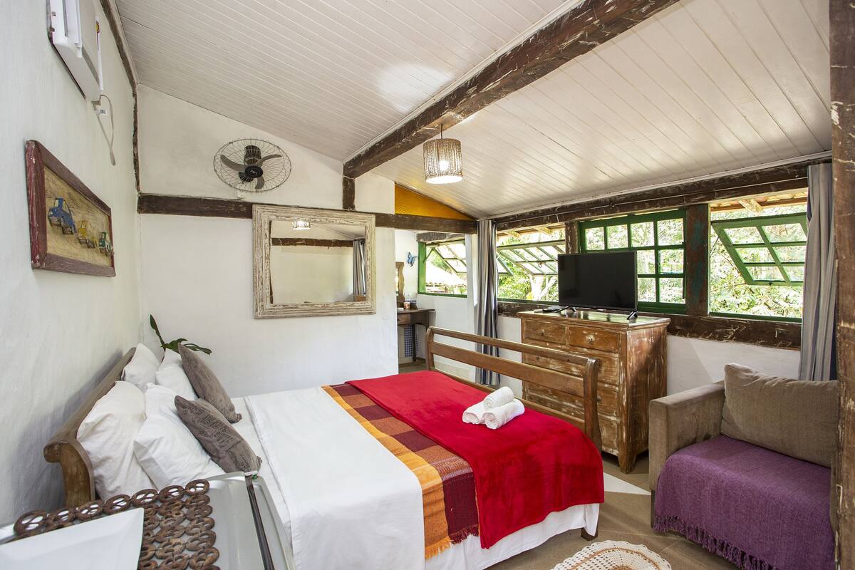 Une chambre avec un lit, une commode en bois, un téléviseur et une fenêtre donnant sur un paysage verdoyant.