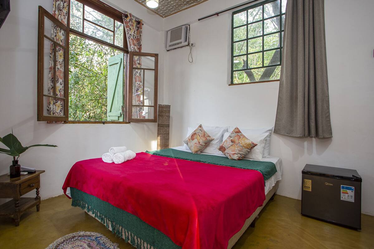 Une chambre avec un lit, un petit réfrigérateur, une fenêtre avec des rideaux et une plante.