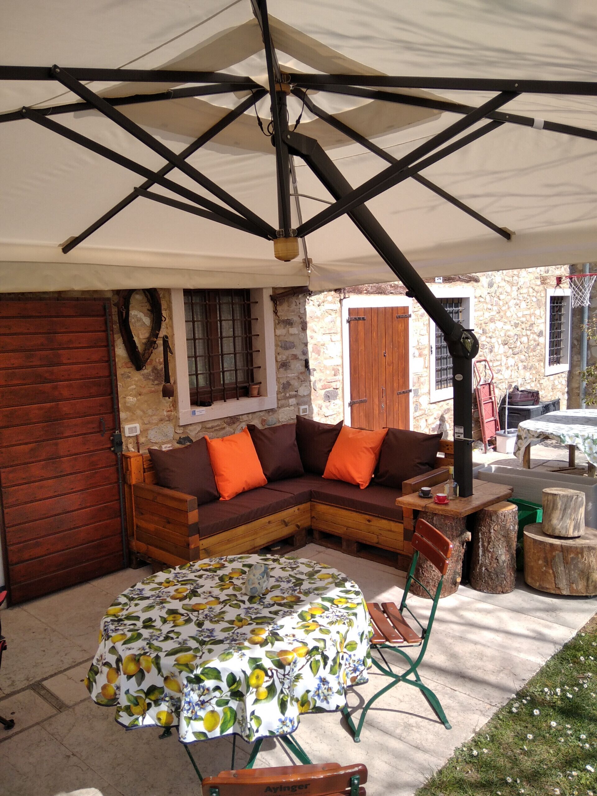 A covered outdoor seating area with a wooden bench, a round table with a floral tablecloth, and a stone wall in the background.