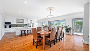 A spacious kitchen with white cabinets, a wooden dining table, and a large window offering a view of the outdoors. - Nelson Bay CBD Oasis - 13 Government Rd (Nelson Bay)