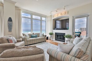 Un salón amplio con grandes ventanales con vistas al océano, una chimenea y un letrero que dice 'BEACH HOUSE'.