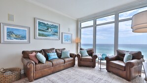 Ett vardagsrum med ett stort fönster med utsikt över havet, inrett med en brun lädersoffa och fåtöljer, ett litet sidobord och inramade konstverk med havstema på väggen.