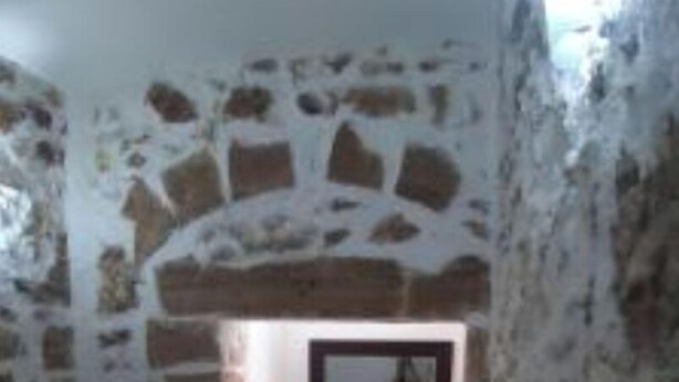 Un couloir étroit, surmonté d’une arche en pierre, mène à une pièce. Les murs sont faits de pierres de formes irrégulières et le sol est carrelé.
