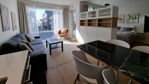 En moderne stue med sofa, spisebord og stole. Stort vindue med gardiner, udsigt til gaden og en balkon.